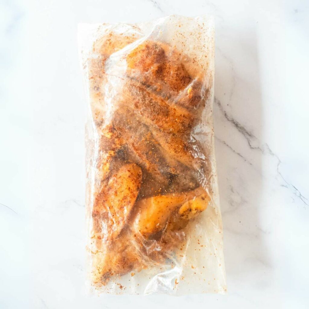 Seasoned chicken wings in a clear plastic bag on a marble surface.