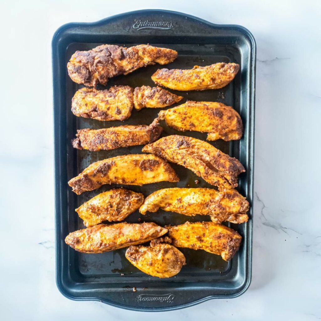 Baked, seasoned chicken tenderloins arranged in two rows on a black baking tray.