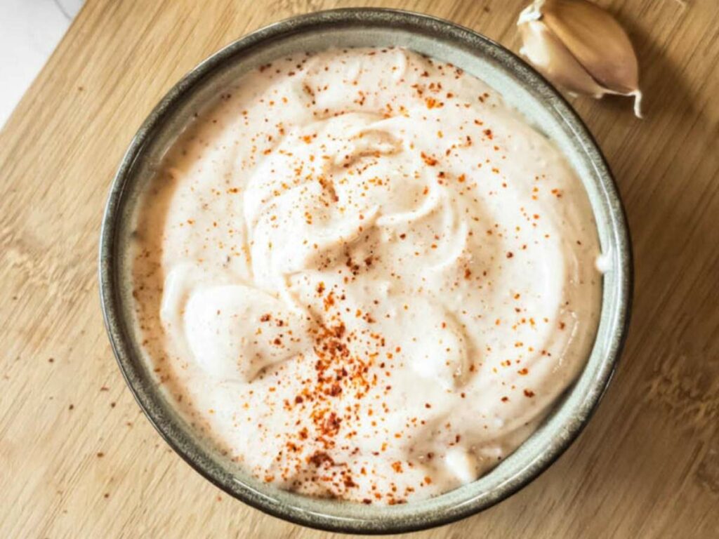 A bowl of chipotle mayonnaise, topped with a sprinkle of paprika, sits on a wooden surface next to a garlic clove.