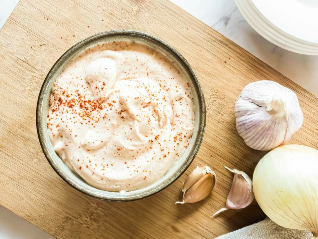 A bowl of chipotle mayonnaise sprinkled with paprika sits on a wooden board next to two garlic bulbs, garlic cloves, and an onion.