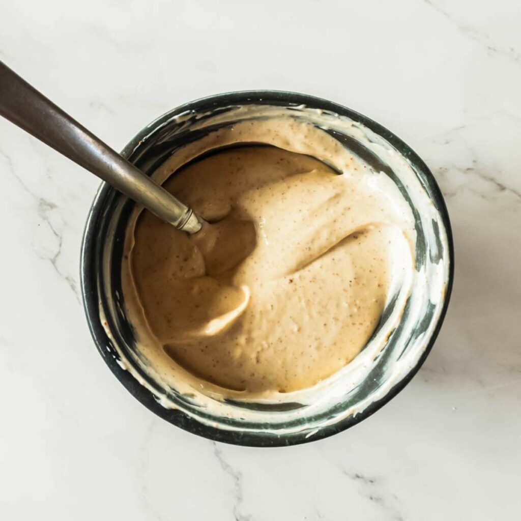 Bowl filled with creamy, beige sauce or dressing, stirred with a metal spoon, placed on a light marble surface.