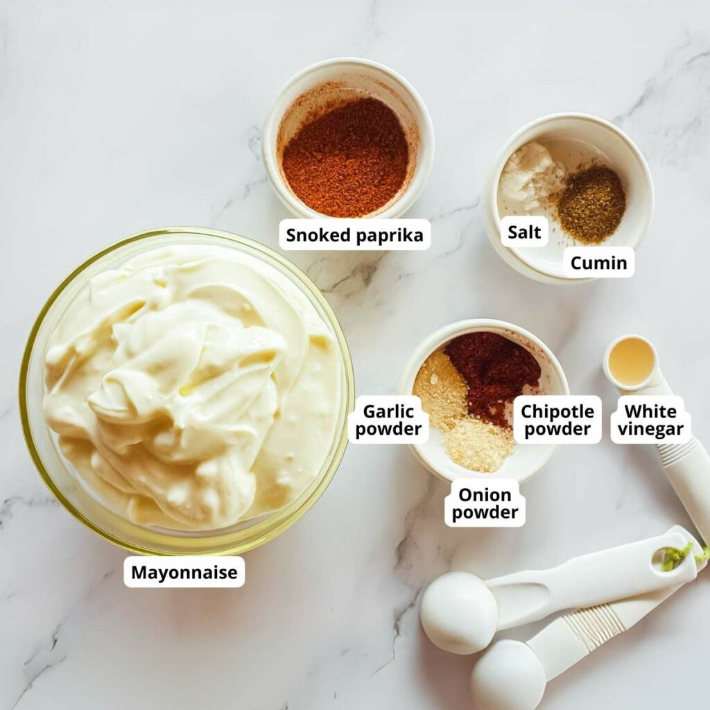 Ingredients for a chipotle mayonnaise sauce on a marble surface include mayonnaise, smoked paprika, chipotle powder, garlic powder, onion powder, cumin, salt, and white vinegar. Measuring spoons nearby.