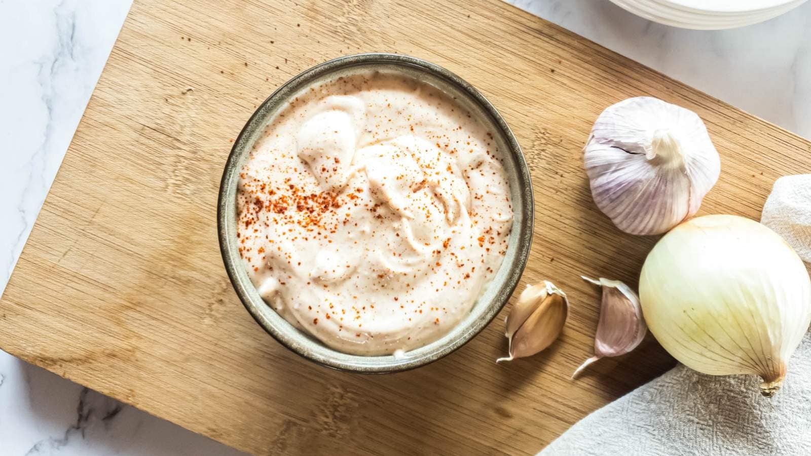 Bowl of chipotle mayonnaise with sprinkled paprika on a wooden board, surrounded by garlic and an onion.