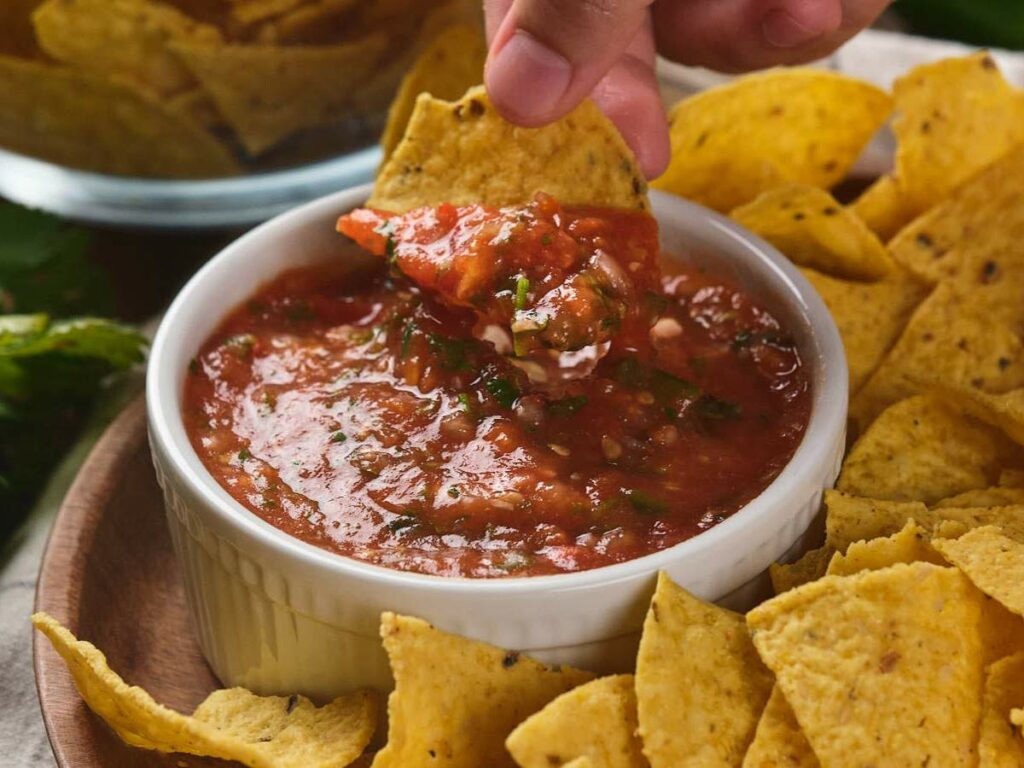 Hand dipping a tortilla chip into a bowl of red salsa surrounded by more chips.