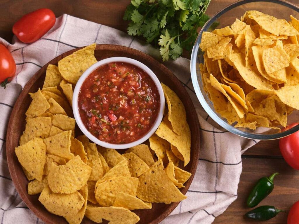 Bowl of salsa in the center of a wooden plate surrounded by tortilla chips, with additional chips.