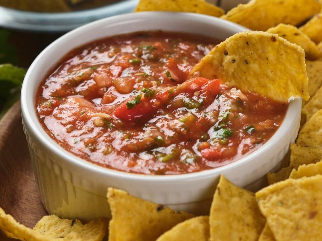 A bowl of tomato salsa with a tortilla chip dipped in it, surrounded by more chips.