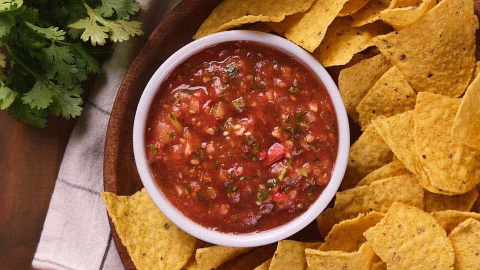 A white bowl of salsa surrounded by tortilla chips on a wooden platter, with a sprig of cilantro nearby.