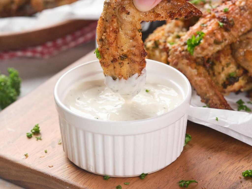 A chicken wing being dipped into ranch sauce in a small ramekin, with more wings and parsley garnish in the background.