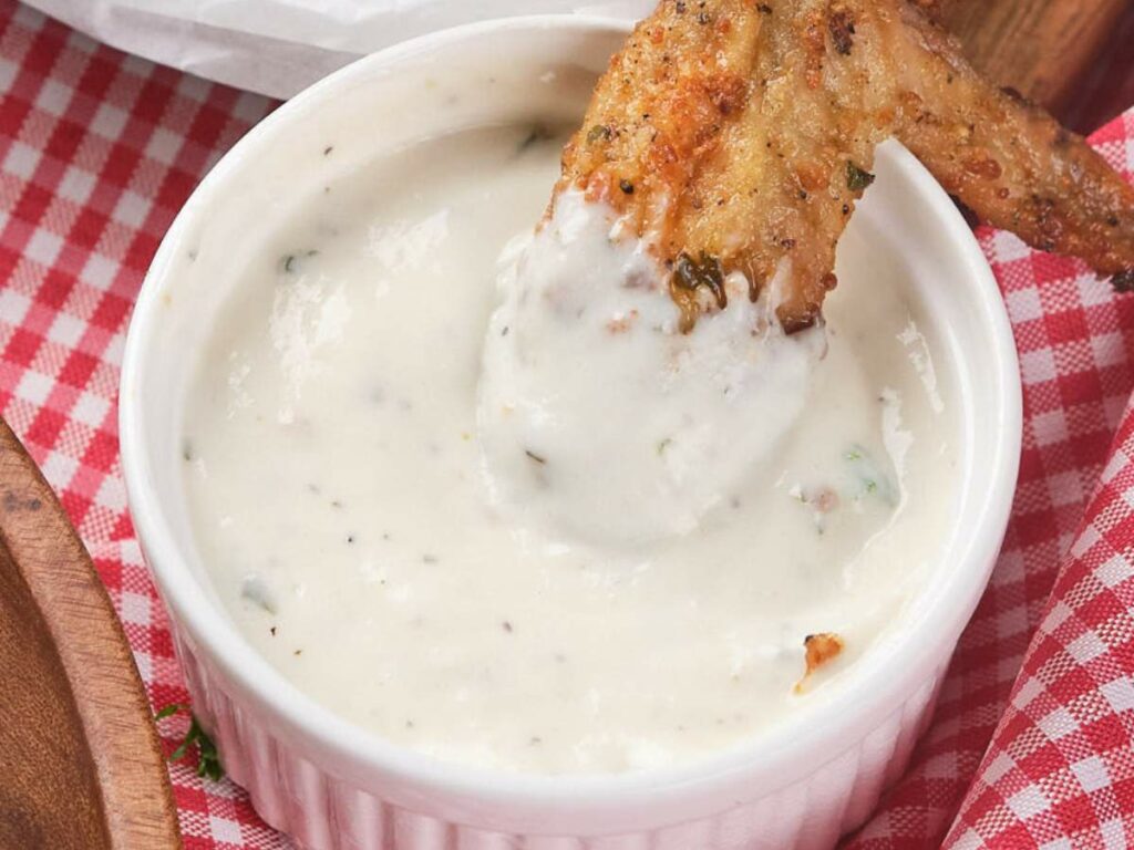 A chicken wing is being dipped into ranch sauce in a ramekin, placed on a red checkered cloth.