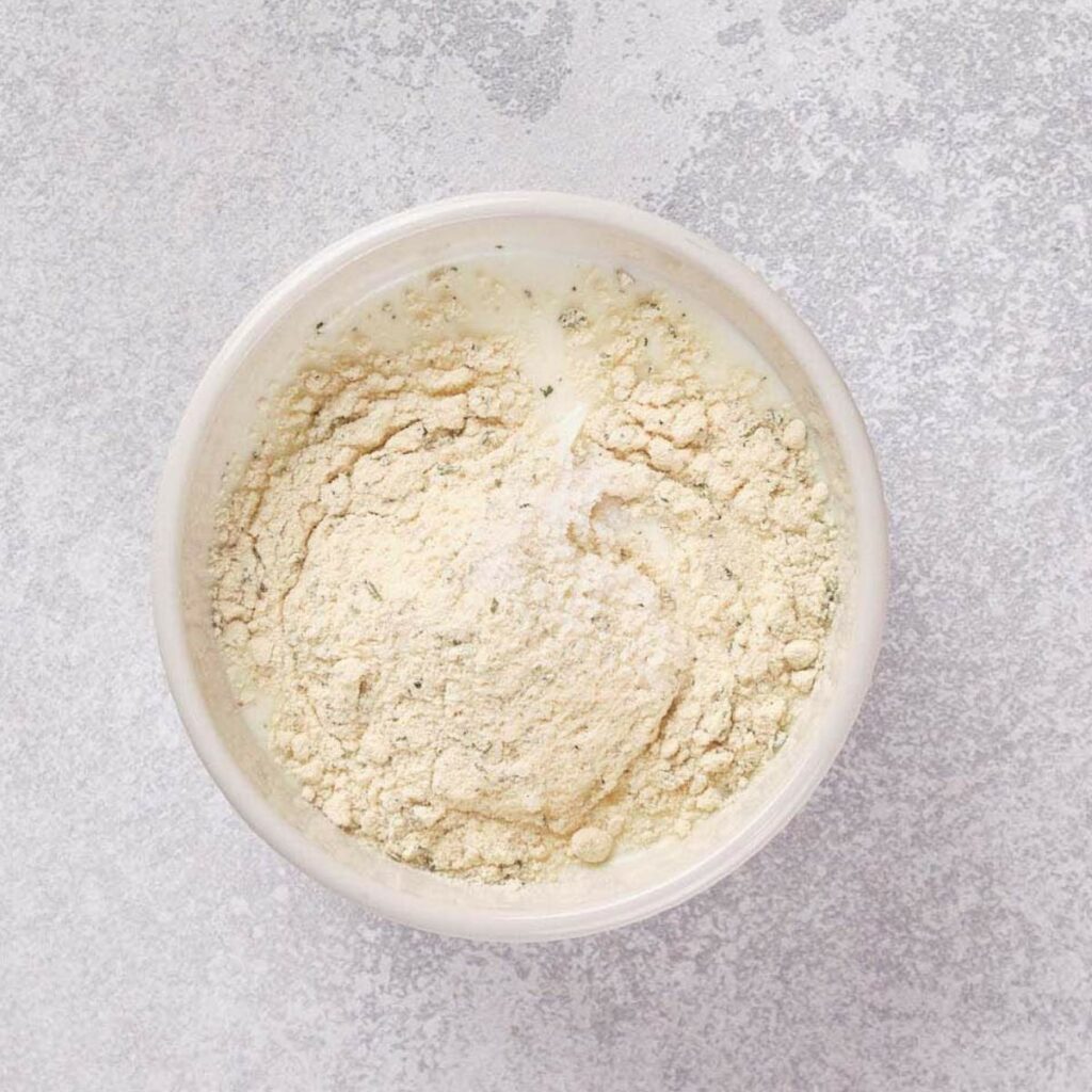 A bowl containing white powder mixture, possibly a seasoning blend or baking ingredient, on a textured gray surface.