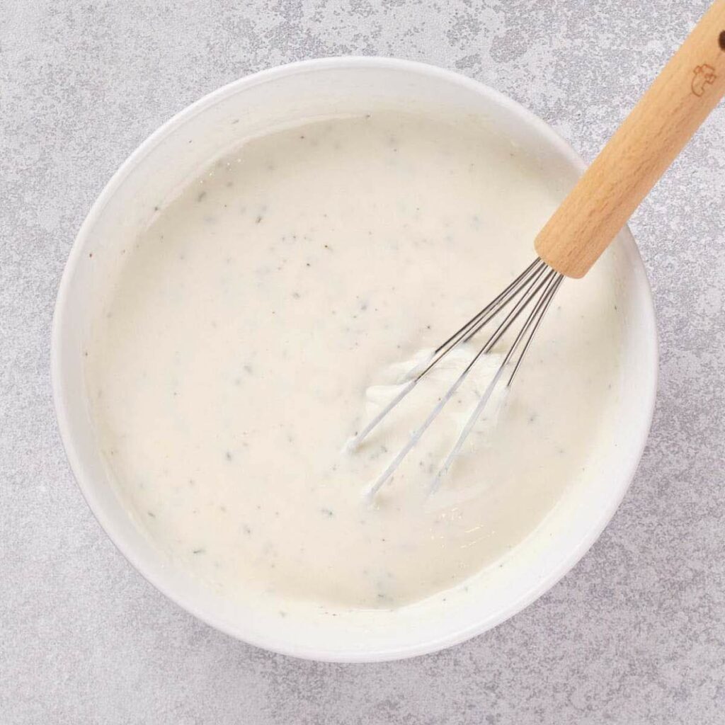 A bowl of creamy white sauce with scattered herbs, being mixed with a wooden-handled whisk on a textured gray surface.