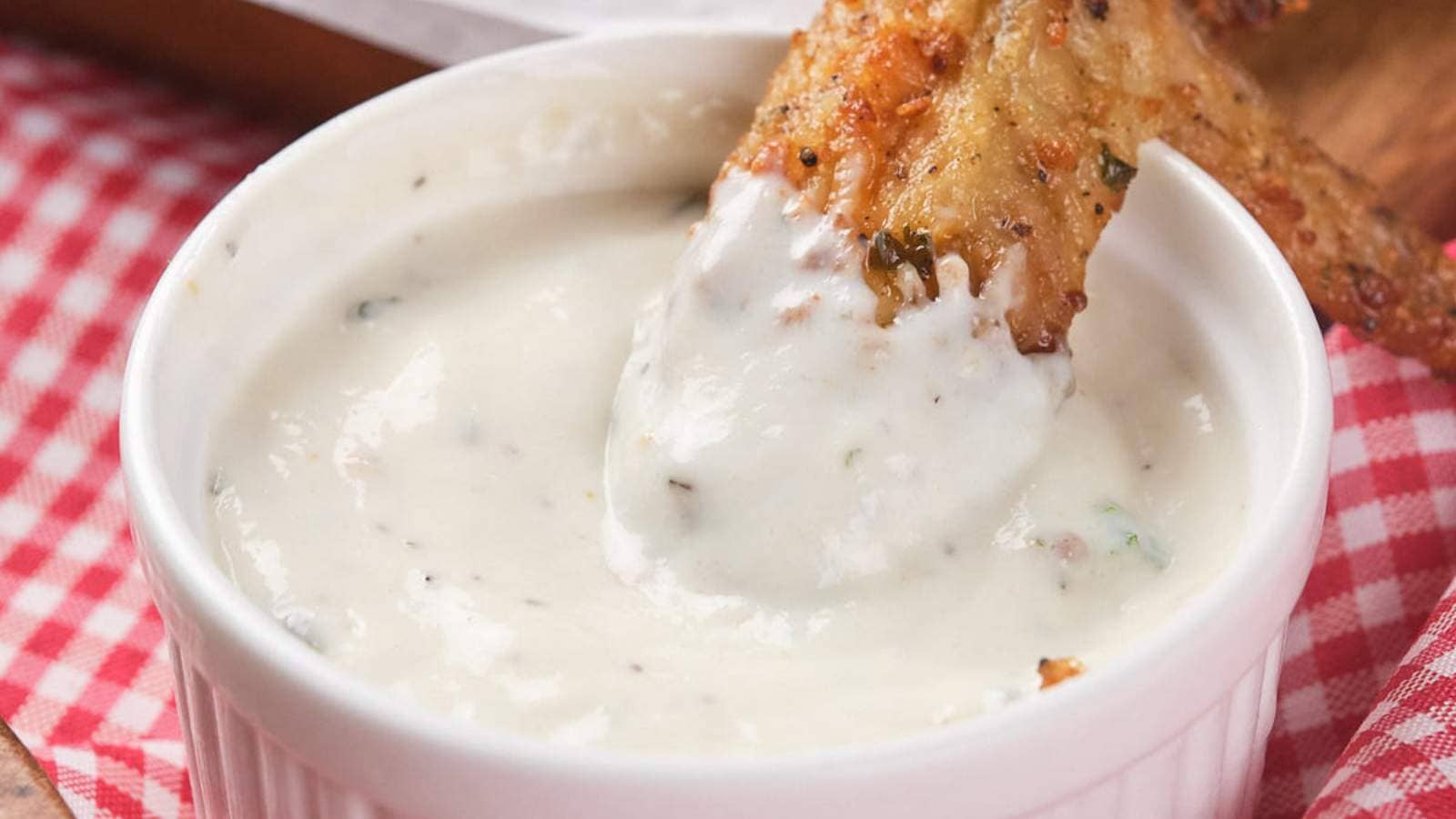 Chicken wing dipped in a ranch sauce in a small white bowl, placed on a red and white checkered cloth.