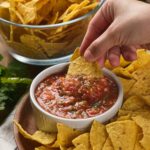 A hand dips a tortilla chip into a bowl of salsa, surrounded by more chips.