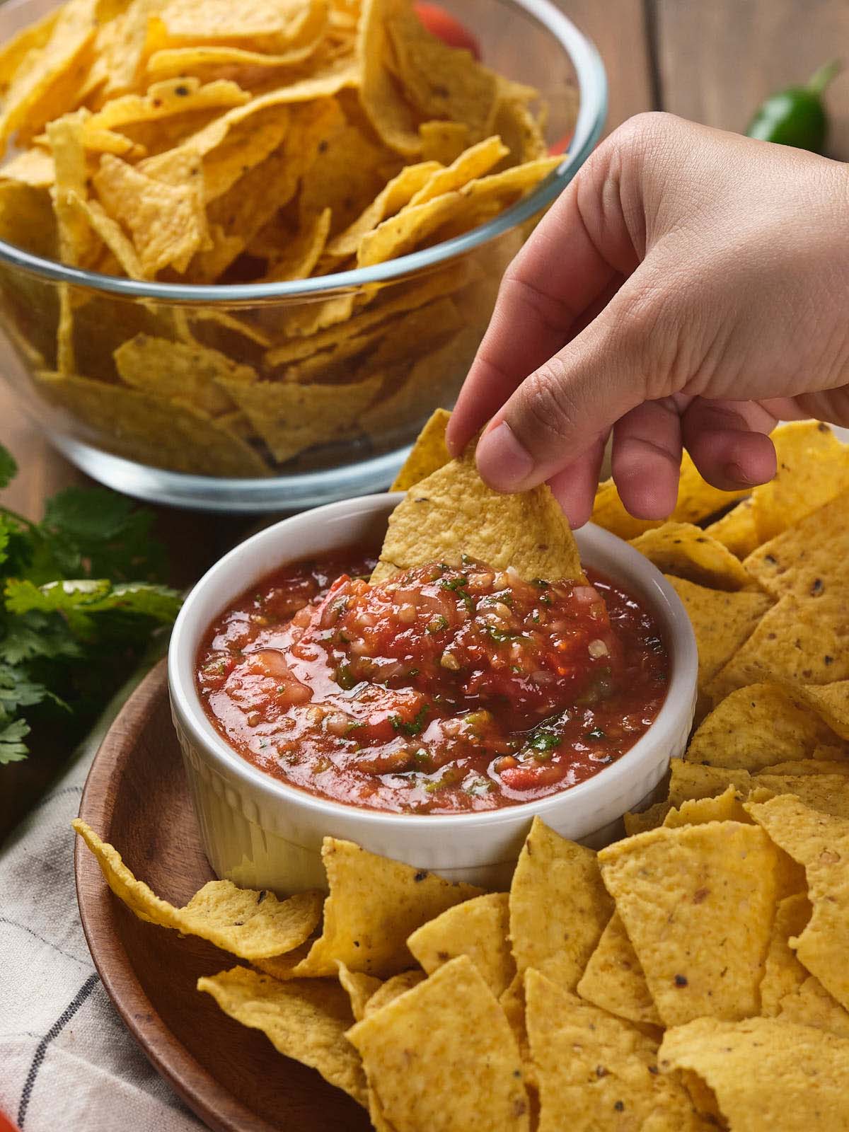 A hand dips a tortilla chip into a bowl of salsa, surrounded by more chips.