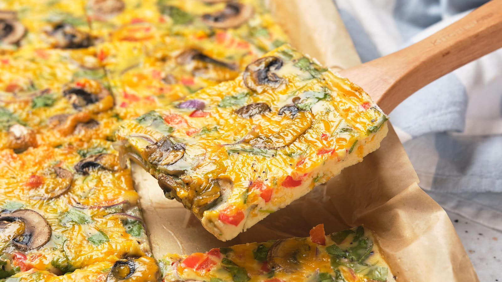 A square slice of sheet pan omelet on a wooden spatula above parchment paper.