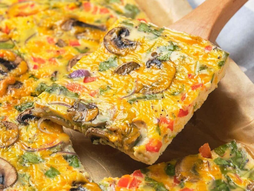 A slice of sheet pan omelet with mushrooms, bell peppers, and herbs on a wooden spatula.