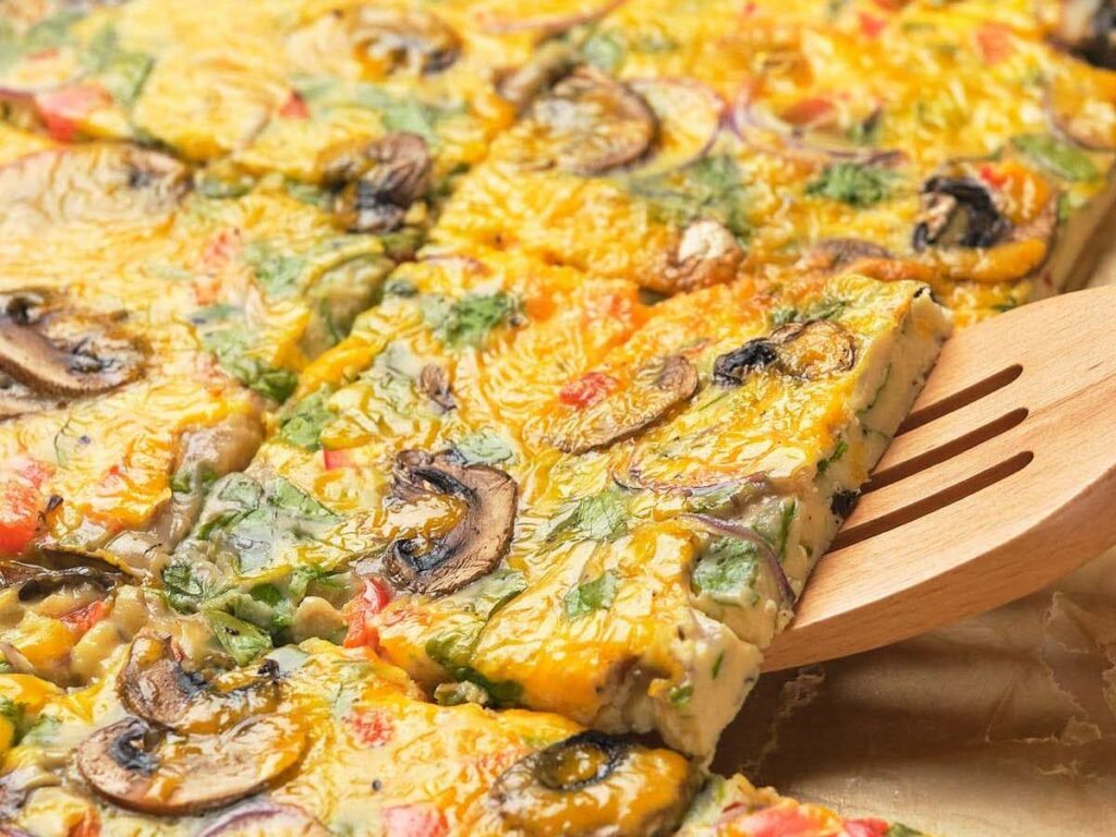 Close-up of sheet pan omelet with mushrooms, spinach, and tomatoes, cut into square pieces.