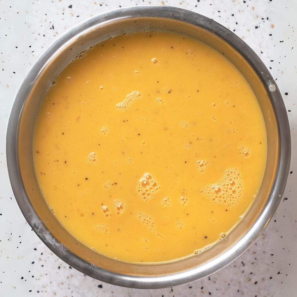 A stainless steel bowl filled with smooth, yellow batter sits on a speckled white countertop.