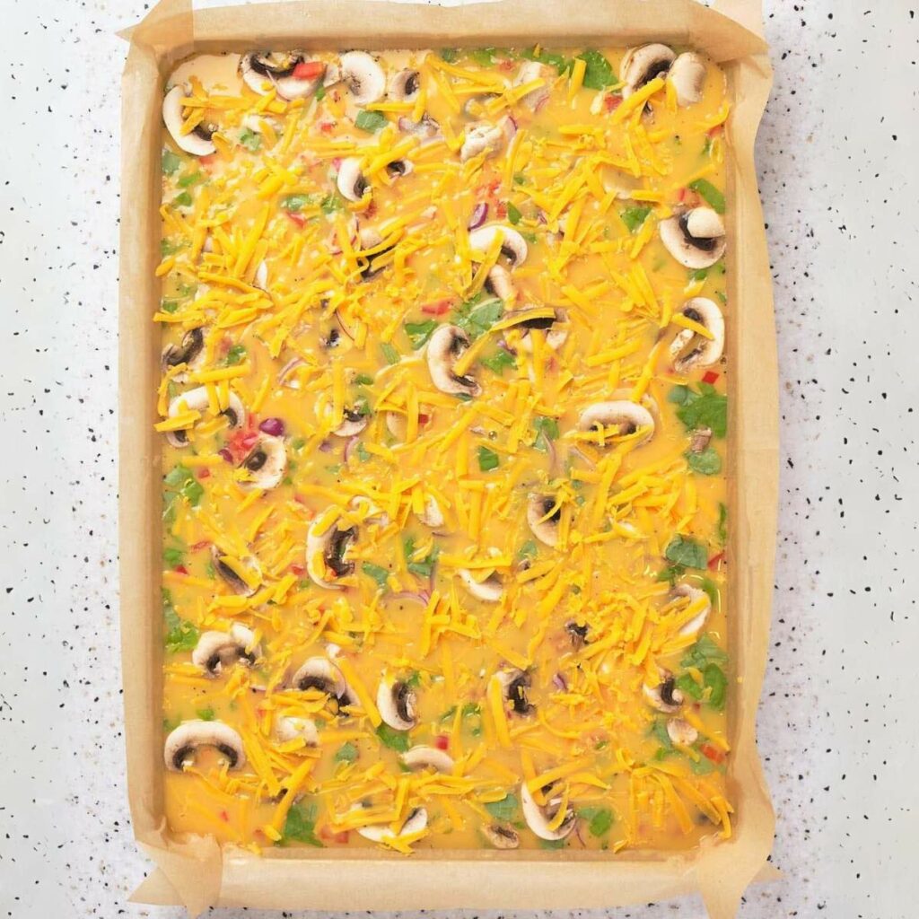 Unbaked egg mixture on a parchment-lined baking sheet with chopped vegetables, mushrooms, and shredded cheese on a speckled countertop.