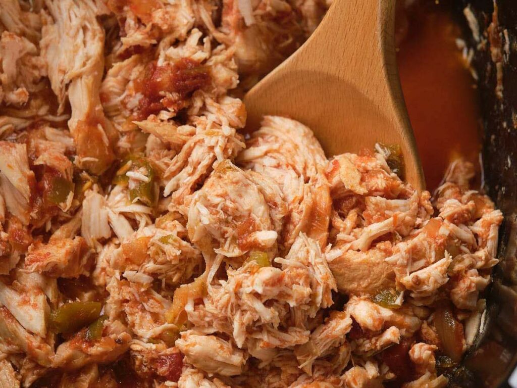 Close-up of shredded salsa chicken mixed with sauce and vegetables, being stirred with a wooden spoon.