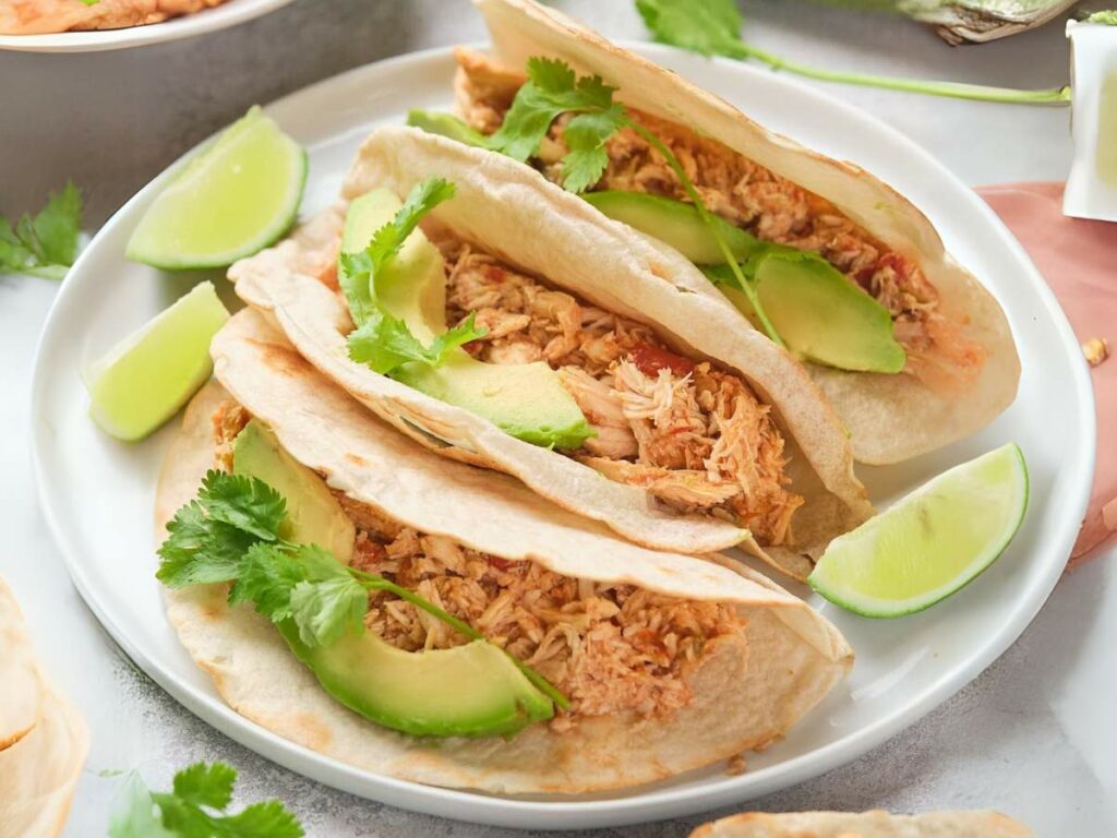 Three soft tacos with shredded chicken, avocado slices, and cilantro on a plate, garnished with lime wedges.