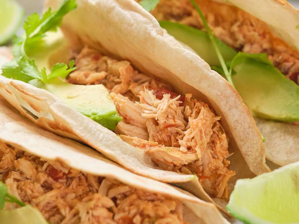 Close-up of tacos with shredded chicken, avocado slices, and cilantro, served with lime wedges.