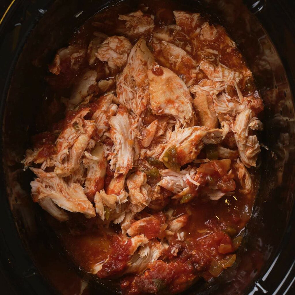 Shredded chicken in a tomato-based sauce with visible chunks of vegetables in a slow cooker.