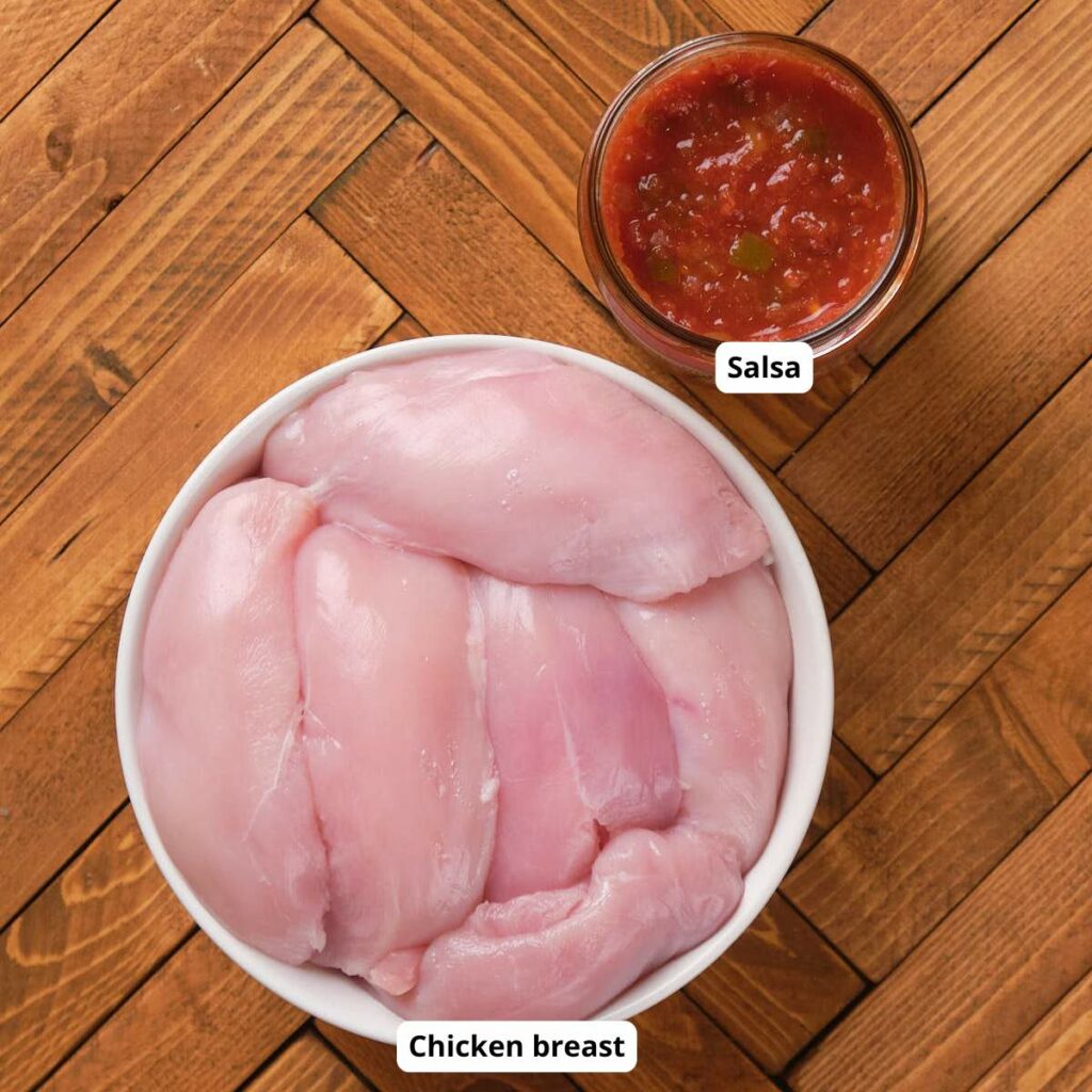 A bowl of raw chicken breasts is on a wooden surface next to a small glass bowl of salsa.