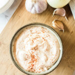 A bowl of chipotle mayonnaise sprinkled with paprika on a wooden board, surrounded by garlic bulbs, cloves, and an onion.