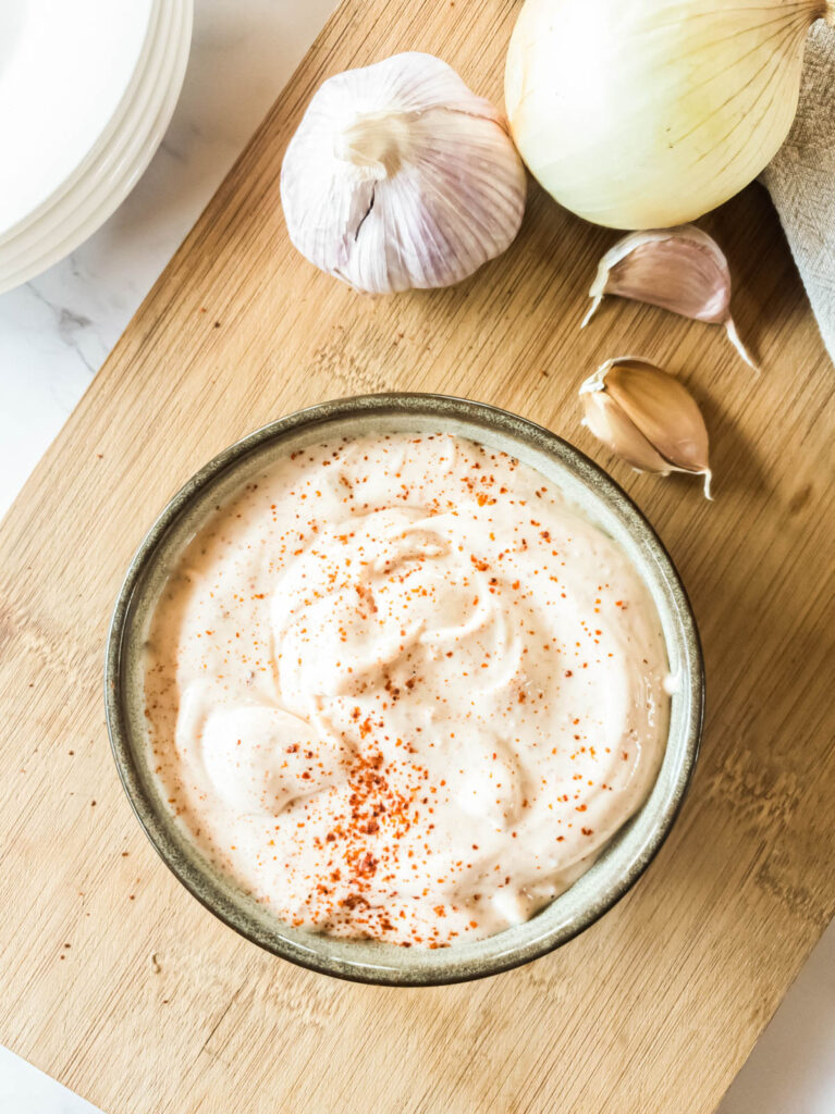 A bowl of chipotle mayonnaise sprinkled with paprika on a wooden board, surrounded by garlic bulbs, cloves, and an onion.