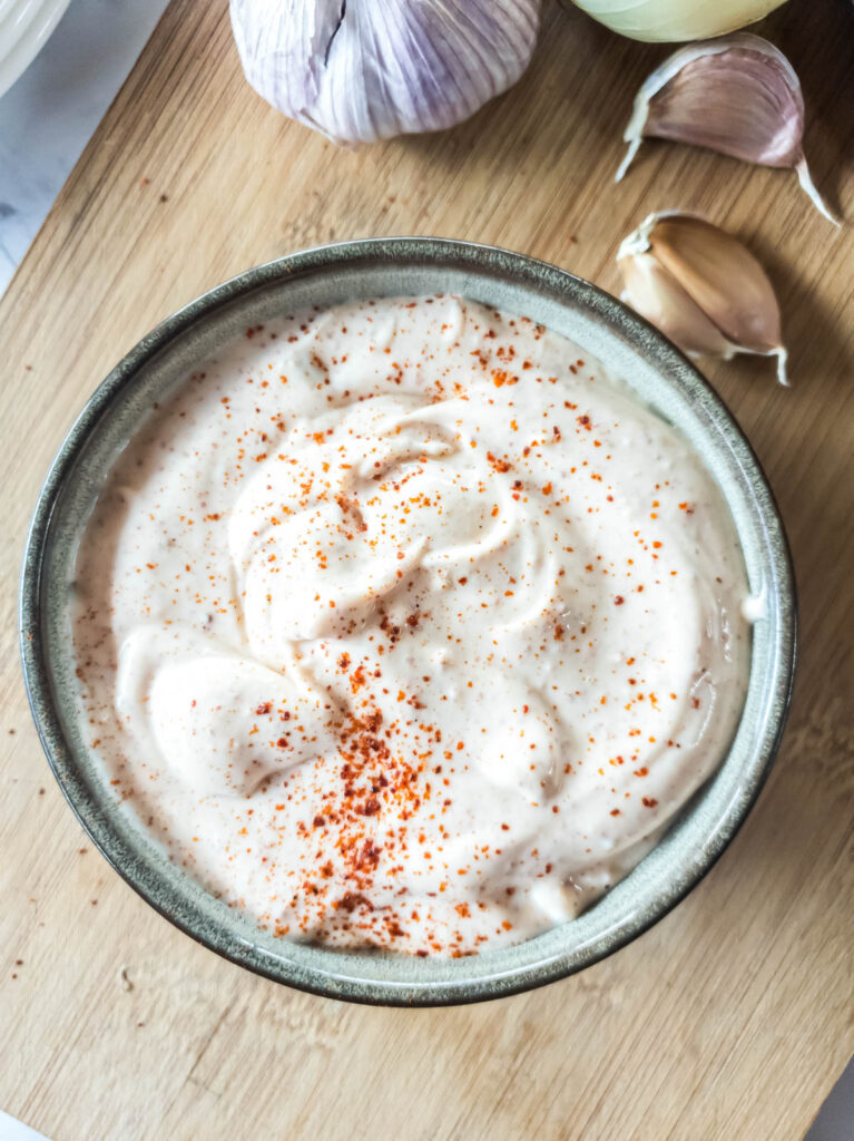 Bowl of chipotle mayonnaise sprinkled with paprika on a wooden surface, surrounded by garlic cloves and bulbs.