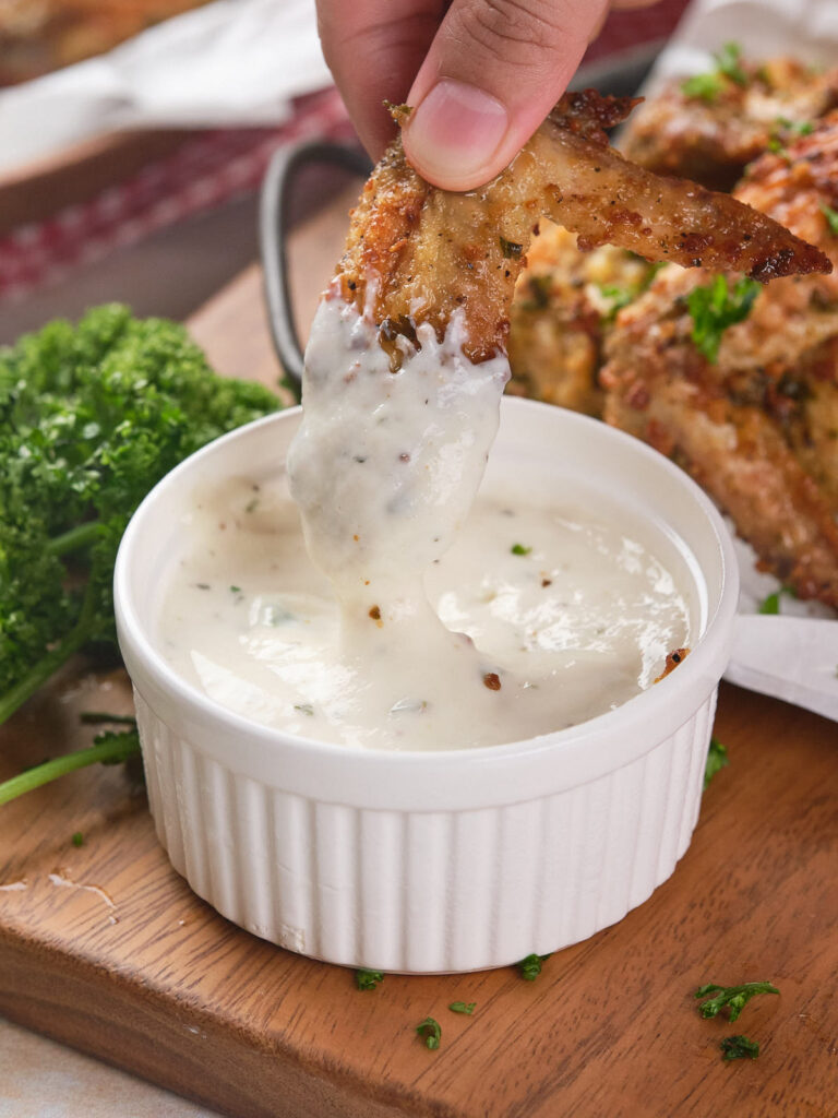 A chicken wing is being dipped into ranch sauce in a small ramekin. Parsley is visible on the wooden surface.