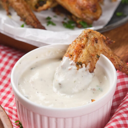 A chicken wing is being dipped into a ranch sauce in a small ramekin.