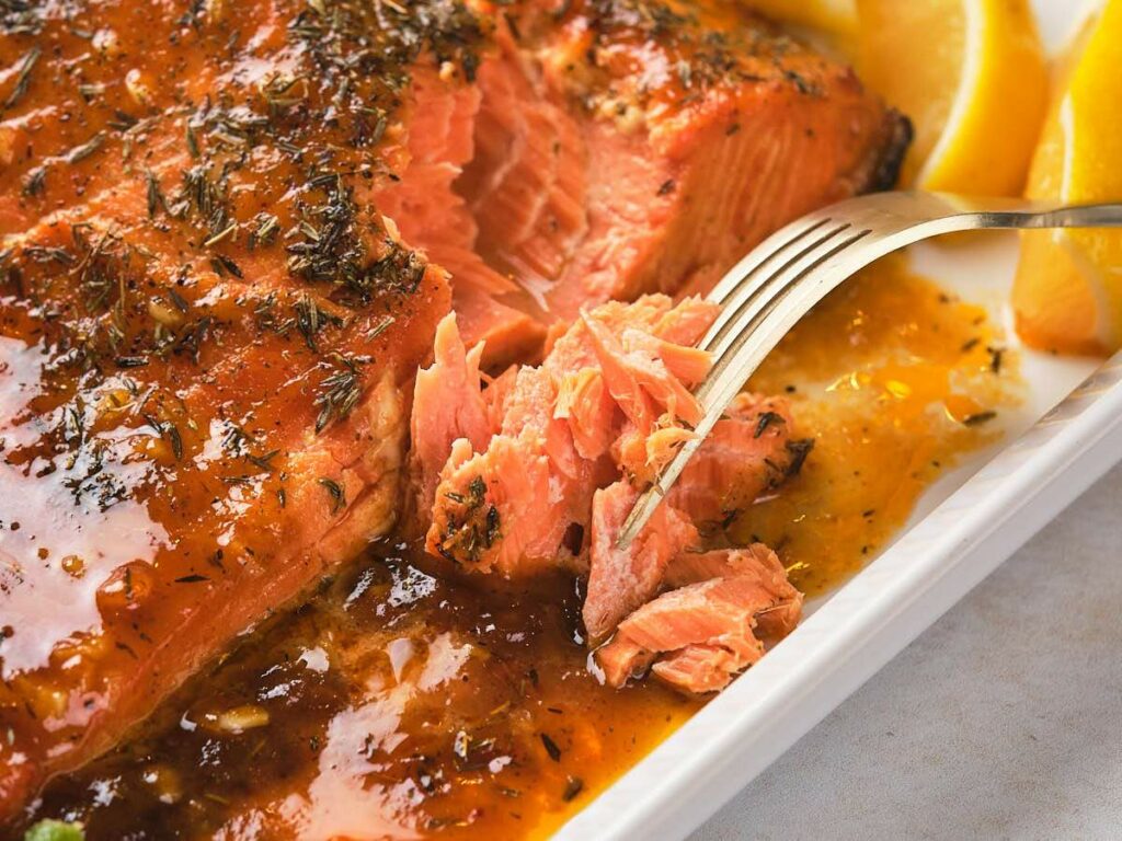 Fork breaking into a glazed, baked salmon fillet garnished with herbs and accompanied by lemon wedges on a white plate.