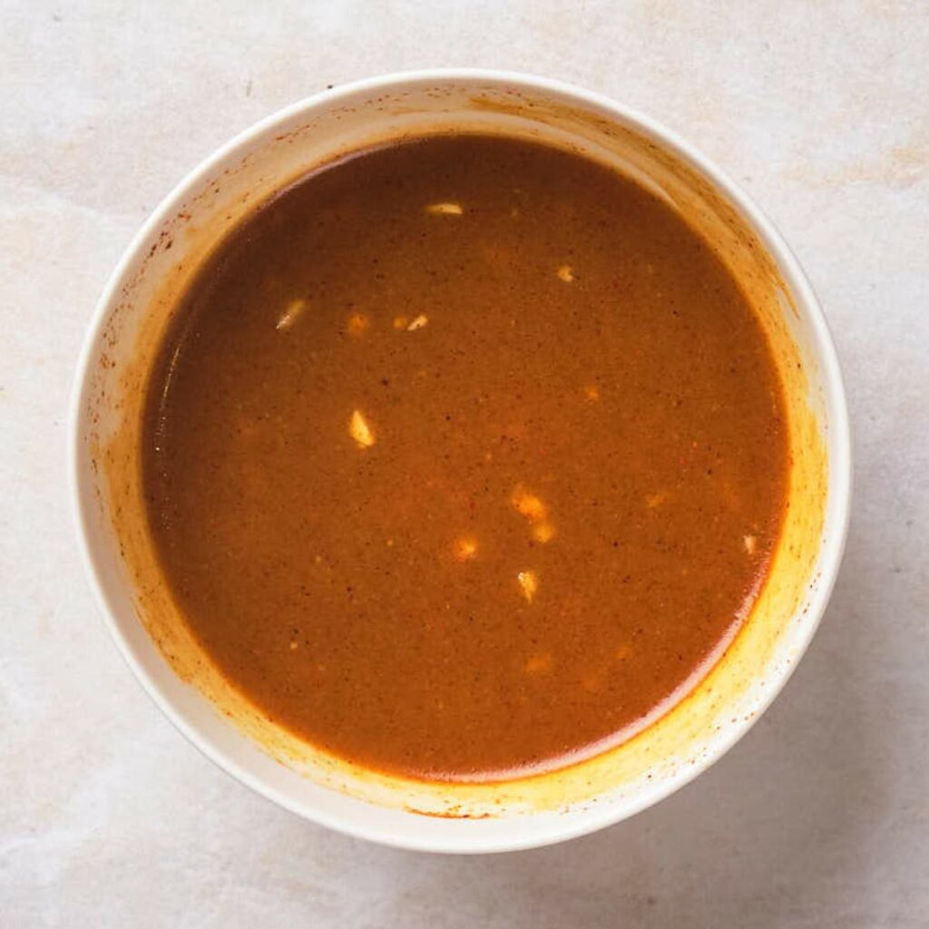 A white bowl filled with brown enchilada sauce, showing small bits of spices and garlic, placed on a light-colored surface.