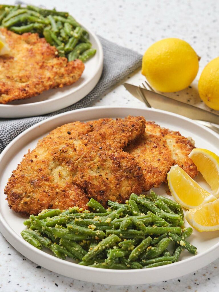 Plates of breaded fried chicken cutlets with seasoned green beans and lemon wedges. Two whole lemons are visible in the background.