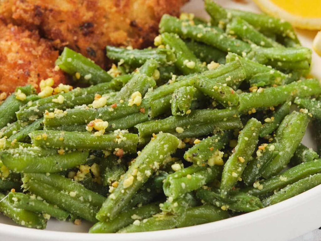 A plate of cooked green beans seasoned with breadcrumbs and herbs, with a hint of lemon in the background.