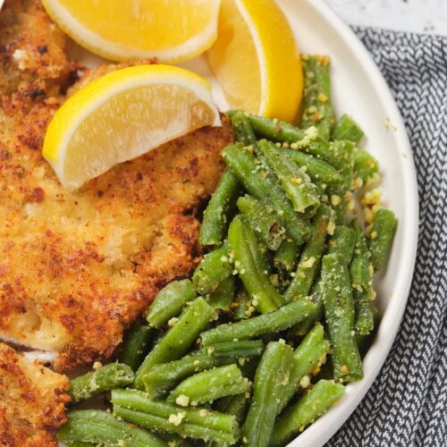 A plate with breaded chicken cutlets, green beans with seasoning, and lemon wedges, on a gray patterned napkin.