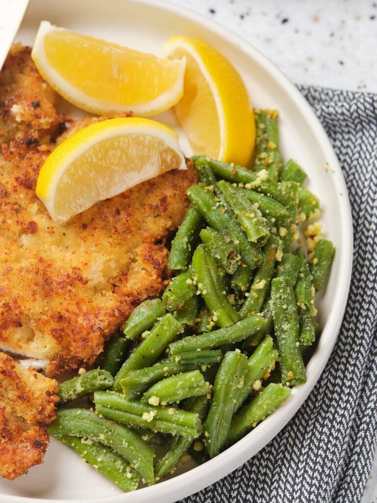 A plate with breaded chicken cutlets, green beans with seasoning, and lemon wedges, on a gray patterned napkin.