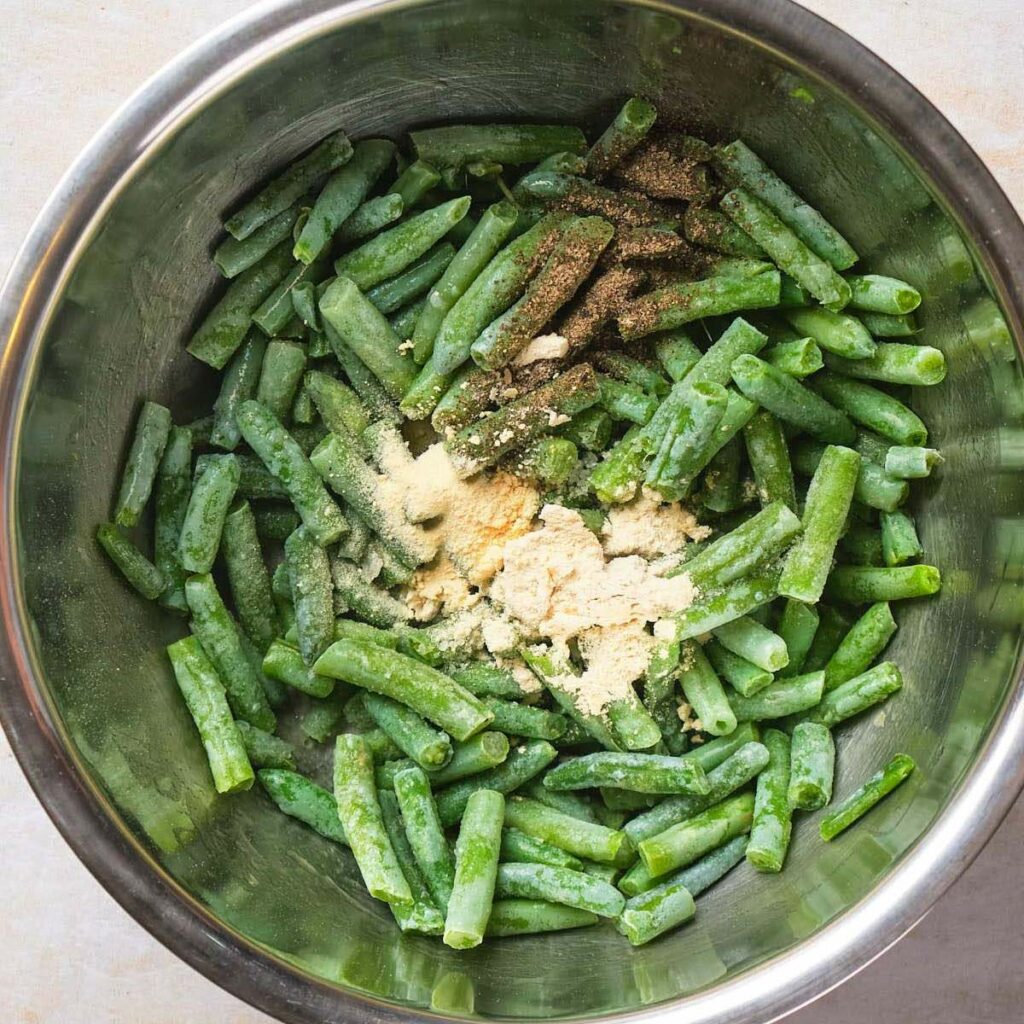 A metal bowl containing frozen green beans, sprinkled with seasoning and powdered ingredients.