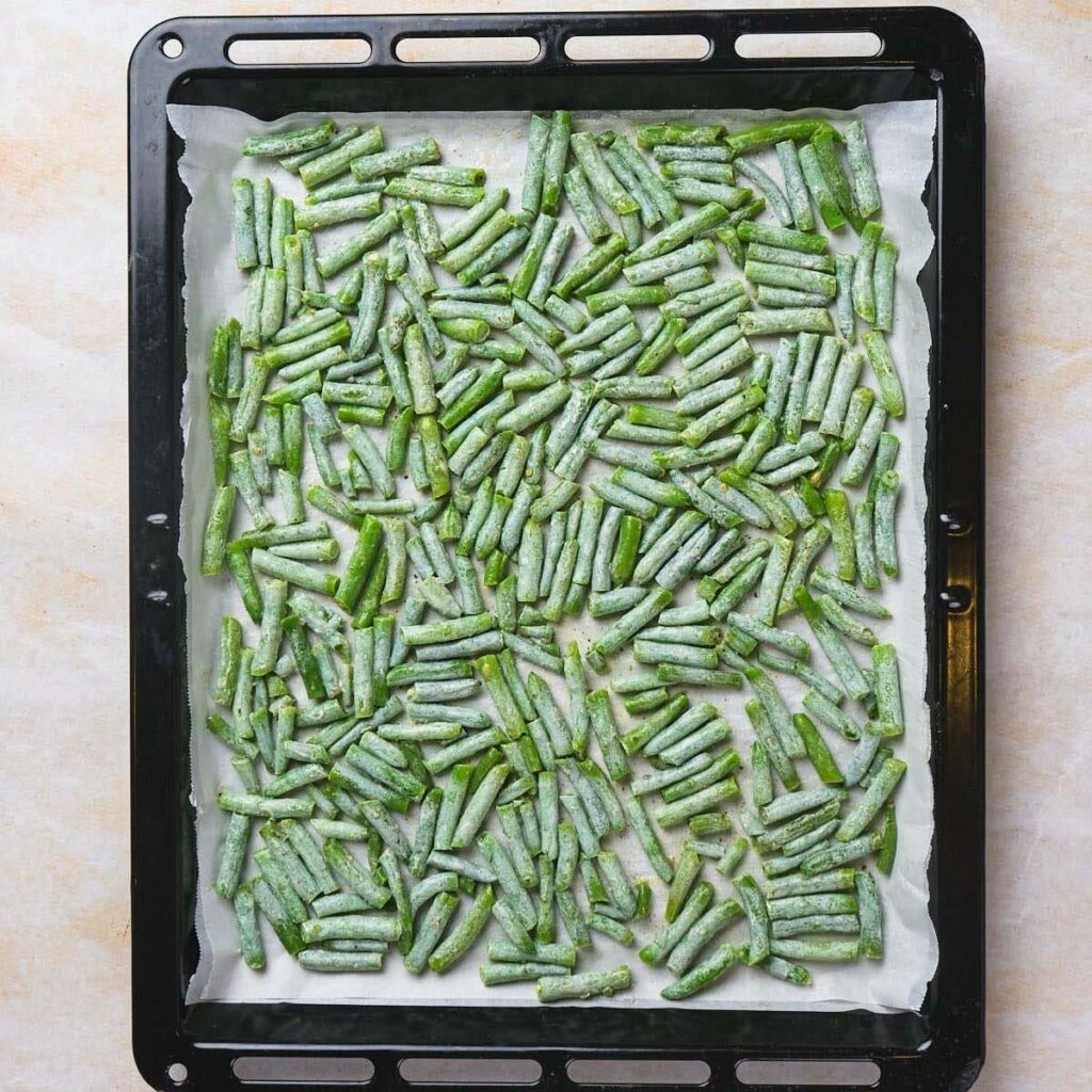 Frozen green beans spread evenly on a parchment-lined baking tray.