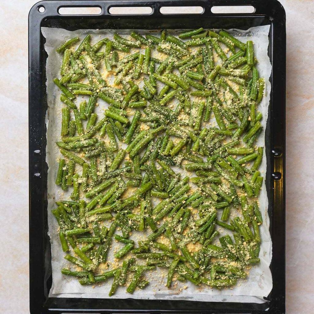 Green beans coated with seasoning and cheese on a parchment-lined baking tray.